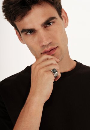 Srebrny pierścionek z teksturowanym czarnym wzorem siatki, noszony na palcu wskazującym, eksponowany na tle czarnej koszulki i neutralnym tle.