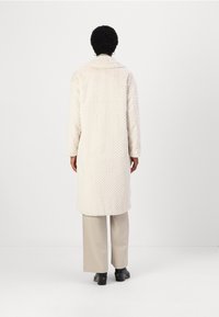 Weißer Mantel im Fischgrätmuster mit einem pelzähnlichen Texture, ausgestattet mit einem breiten Kragen, langen Ärmeln und einem knielangen Schnitt, getragen über beigen Hosen.