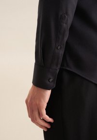 Camicia nera in tessuto testurizzato, con polsini abbottonati e una vestibilità su misura. Primo piano del polso con dettaglio del bottone visibile.