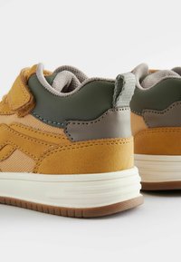 Tan en groene sneaker met een gestructureerde mesh bovenkant, suede accenten en een gevoerde kraag. Bevat een witte middenzool en rubberen zool.