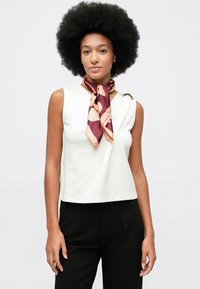 Mulher com grande cabelo afro usa uma blusa branca sem mangas, calças pretas e um lenço padrão em marrom e rosa amarrado ao redor do pescoço.