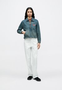 Γυναίκα στέκεται μπροστά σε λευκό φόντο, φορώντας μπλε τζιν μπουφάν, καφέ πουκάμισο, λευκό παντελόνι και μαύρα παπούτσια, με το ένα χέρι στην τσέπη του μπουφάν.