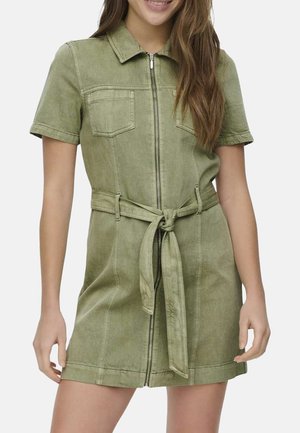 Jeune femme portant une robe ceinturée vert olive à manches courtes avec fermeture éclair à l’avant et poches poitrine, souriante devant un fond uni.