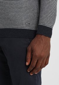 Sweter z dzianiny w poziome szare i czarne paski, z prążkowanym kołnierzem i mankietami. Widoczna metka marki na dolnym brzegu.