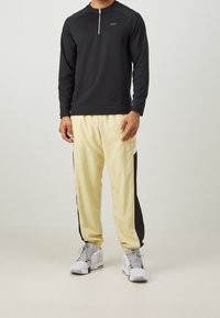 Schwarzes Langarmshirt mit Viertelreißverschluss; beige Jogginghose mit schwarzen Seitenpaneelen; graue Sneaker mit weißen Akzenten; glatte Texturen.