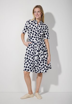 Navyblaues Polka-Dot-Kleid mit kurzen Ärmeln, Kragen und Taillengürtel. Verfügt über zwei seitliche Taschen und einen fließenden Rock. Getragen mit beigen Schuhen.