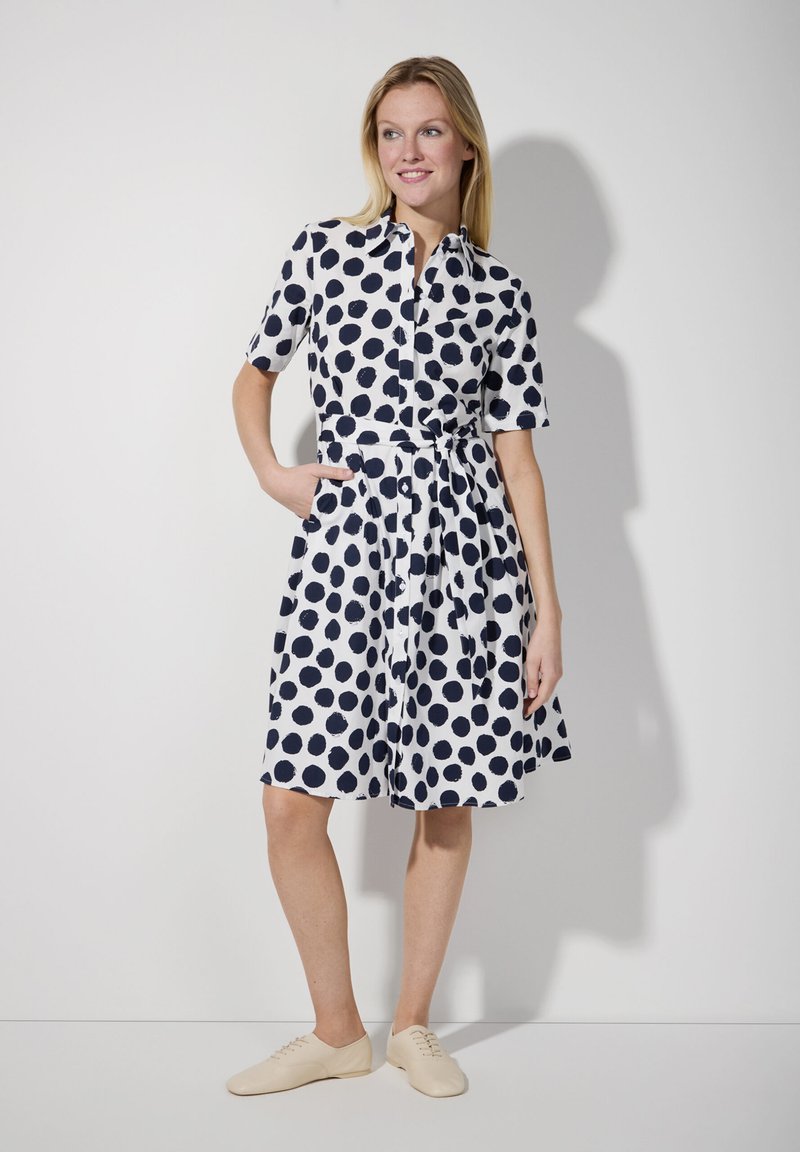 Navyblaues Polka-Dot-Kleid mit kurzen Ärmeln, Kragen und Taillengürtel. Verfügt über zwei seitliche Taschen und einen fließenden Rock. Getragen mit beigen Schuhen.