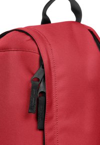 Sac à dos en tissu rouge en gros plan avec des fermetures éclair noires, une surface texturée, des coutures renforcées et une poignée de transport en haut.