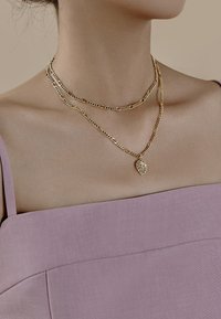 Collares en capas de tono dorado sobre piel desnuda; una cadena presenta un colgante en forma de hoja. El fondo es neutro, con una blusa lavanda visible.