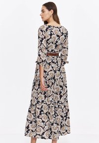 Woman wearing a black paisley-patterned midi dress with 3/4 sleeves tied at cuffs and a brown belt, facing sideways on white background.