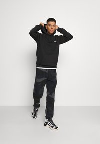 Czarna bluza z kapturem z przednią kieszenią, w połączeniu z ciemnymi spodniami dresowymi w wzór moro. Strój uzupełniają czarno-białe buty sportowe.