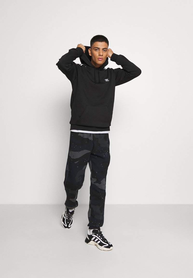 Czarna bluza z kapturem z przednią kieszenią, w połączeniu z ciemnymi spodniami dresowymi w wzór moro. Strój uzupełniają czarno-białe buty sportowe.