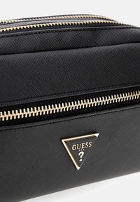 Zwarte textuurstof tas met twee gouden ritsen en een driehoekige Guess USA-logo plaque op het voorvak.