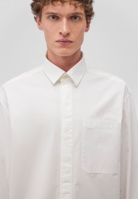 White long-sleeve shirt made of cotton. Button-down collar, front pocket, straight cut, and smooth texture with minimalistic design.