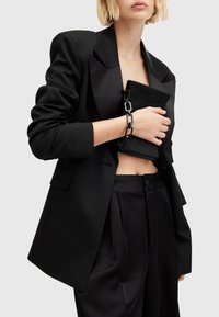 Mustaplasti, jossa on satiinikaulukset, löysä istuvuus, yhdistettynä mustiin satiinipö trousers. Pitelee mustaa laukkua, jossa on ketjuhand. 