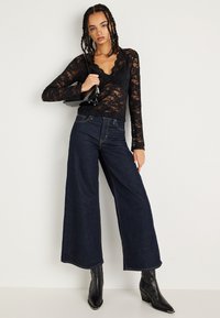 Zwarte kant lange mouw top met gekartelde randen, gecombineerd met hoge taille wijdvallende donkerdenim jeans en puntige zwarte haklaarzen.