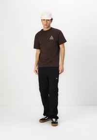 Camiseta de algodón marrón con un logo triangular, combinada con pantalones cargo negros con bolsillos y zapatillas negras con suela marrón.