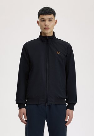 Junger Mann steht aufrecht und trägt eine schwarze Reißverschlussjacke mit einem kleinen, goldfarbenen Sticklogo auf der Brust und dunkle Hosen.