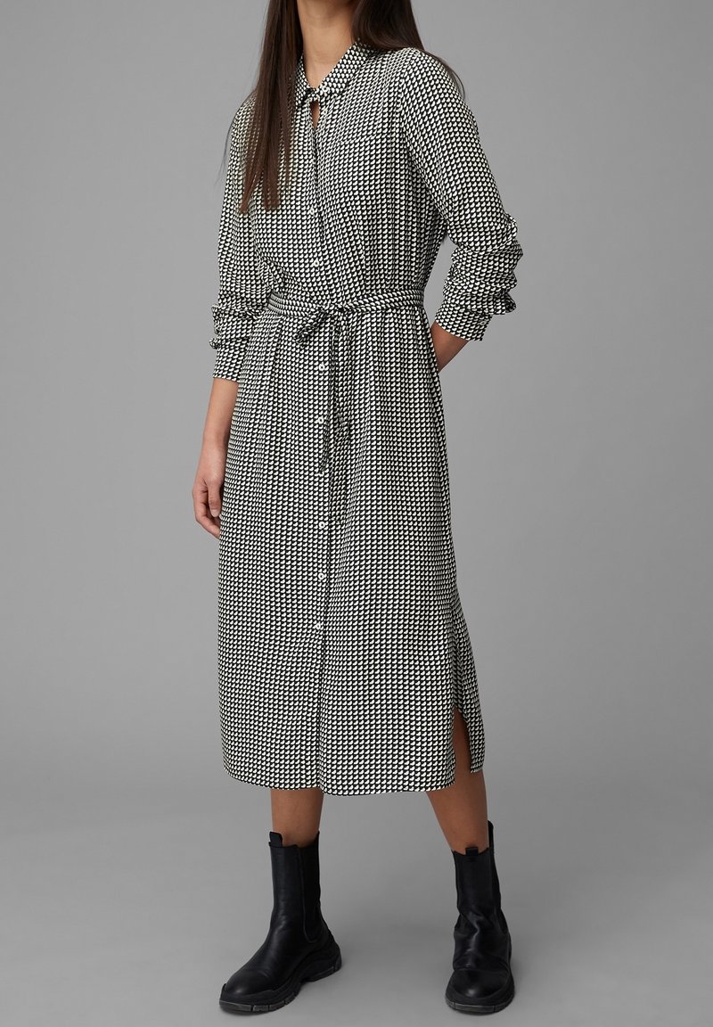 Robe à carreaux noir et blanc avec un devant à boutons, des manches longues, une ceinture à la taille, des fentes sur les côtés et un col, associée à des bottines noires.