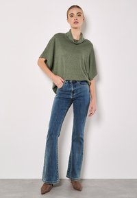 Haut en tricot vert à col bénitier surdimensionné, associé à un jean bleu évasé et des bottes à talons taupe. Texture lisse, coupe décontractée et style casual.
