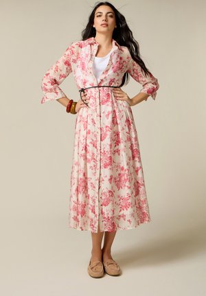 Kvinde med langt mørkt hår iført en hvid midi kjole med rødt blomsterprint, sort bælte, hvid indertop, brune armbånd og beige mokkasiner.