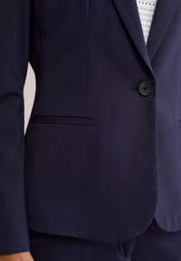 Námořnický modrý saka na míru se zapínáním na jeden černý knoflík, klopovými límci a rozparky, nošené přes bílý perforovaný top.