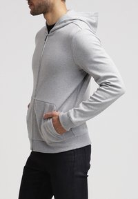 Sudadera con cremallera gris hecha de una tela suave, con un bolsillo frontal, mangas largas y capucha. Diseño minimalista sin patrones visibles ni acentos.