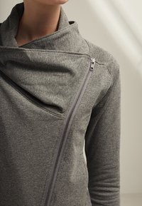 Šedá mikina s volným límcem, s diagonálním zipem a měkkou, texturovanou tkaninou. Detailní švy vylepšují design.