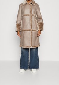 Beige shearling coat with a collared neck, horizontal and vertical seams, side pockets, and plush cuffs paired with wide-leg denim pants.