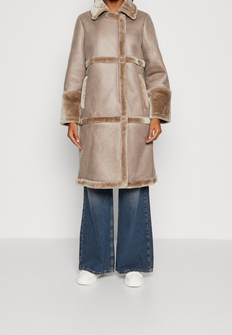 Beige shearling coat with a collared neck, horizontal and vertical seams, side pockets, and plush cuffs paired with wide-leg denim pants.