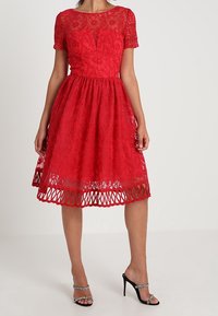 Rotes Spitzenkleid mit kurzen Ärmeln, figurbetontem Oberteil, schwingendem Rock und gewelltem Saum. Tragen mit schwarzen Absatzsandalen und silbernen Akzenten.