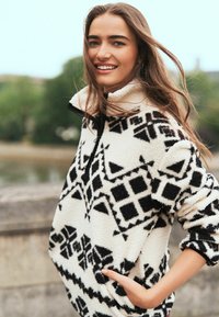 Fleece pullover i creme og sort med geometriske mønstre, lynlås krave og sidelommer. Blød tekstur, afslappet pasform og lange ærmer.
