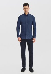 Granatowa koszula z guzikami, w pionowe paski, z długimi rękawami i stożkowym kołnierzykiem, zestawiona z ciemnymi spodniami i czarnymi butami.