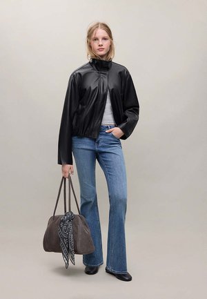 Jeune femme portant une veste en cuir noire, un jean bleu, des bottes noires, tenant un sac à main gris avec un foulard à motif sur un fond uni.