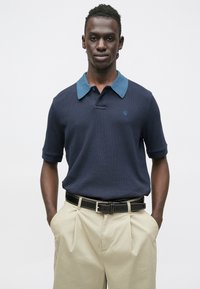 Námornícka polokošeľa s modrým golierom, textúrovaná tkanina, krátke rukávy a malým logom na hrudi; v kombinácii s nohavicami svetlej farby a čiernym opaskom.