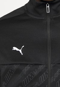 Svart zip-up jacka i slätt tyg, med en vit Puma-logotyp och ett strukturerat mönster med "Motorsport"-tryck på den nedre delen.