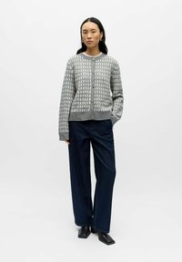 Szary kardigan w wzory z zapięciem na guziki i ściągaczami na rękawach, zestawiony z szerokimi spodniami dżinsowymi w ciemnym kolorze i czarnymi butami o kwadratowych noskach.