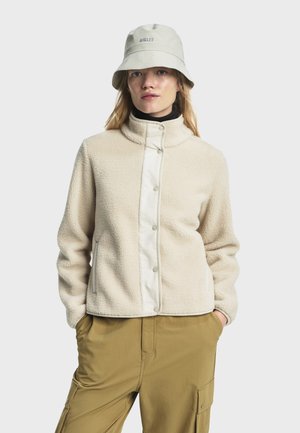 Beige fleecejakke med høj krave, trykknapper og en hvid frontpanel. Båret over en sort top med khaki cargo bukser.