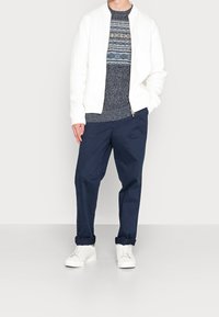 Homme portant une veste blanche à fermeture éclair, un pull à motifs bleu marine, un pantalon bleu marine retroussé et des baskets blanches, debout devant un fond uni.