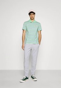 Zaļš polo krekls ar svītrām un apkakli, kopā ar gaiši pelēkām sporta biksēm un zaļām sporta kurpēm. Modelis valkā rozā cepuri.