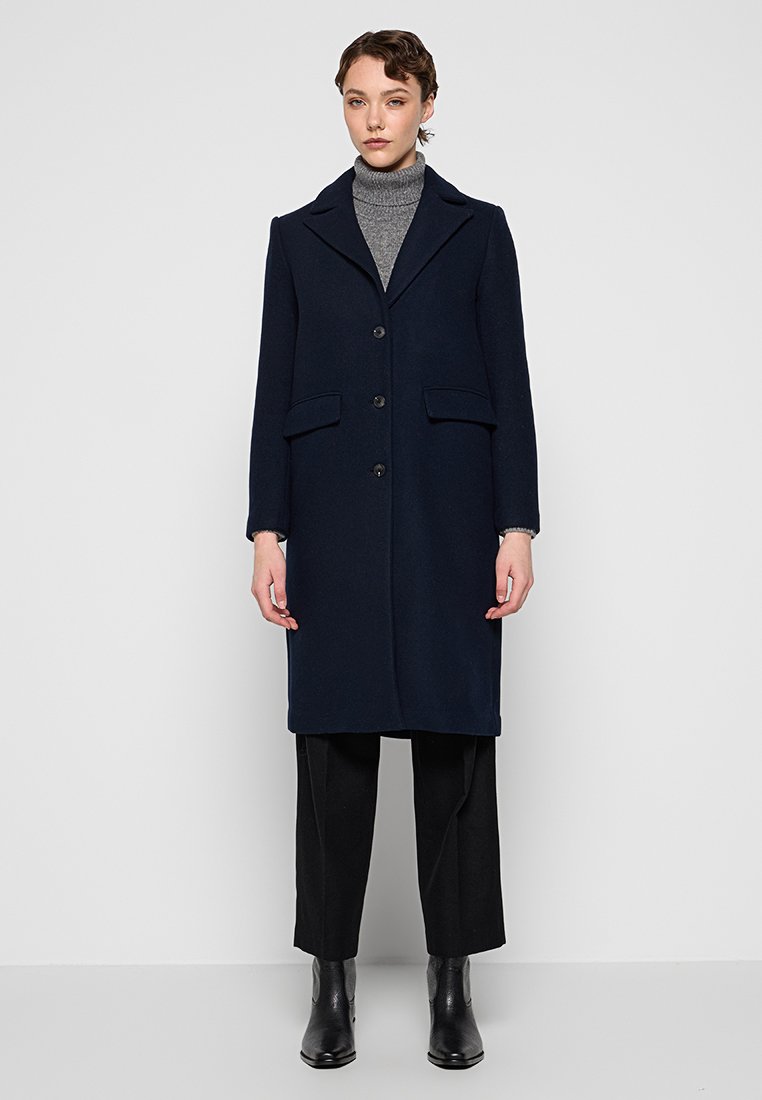 Marineblaue Wollmischung Jacke mit Reverskragen, drei Knöpfen, seitlichen Taschen und langen Ärmeln, kombiniert mit einem grauen Rollkragenpullover und schwarzen Hosen.