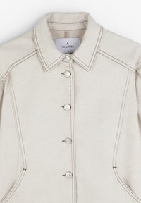 Beige cotton shirt jacket with a pointed collar, six round buttons, contrasting stitching, and side pockets. Label visible inside collar.