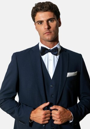 Man buttoning dark navy three-piece suit with white shirt and black bow tie, standing against plain white background.
