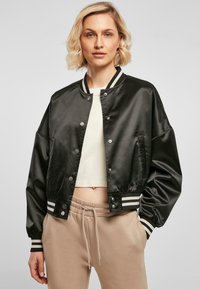 Veste bomber en satin noir avec des poignets et un col rayés, dotée de boutons pression et d'une coupe décontractée. Portée par-dessus un top court et un pantalon beige.