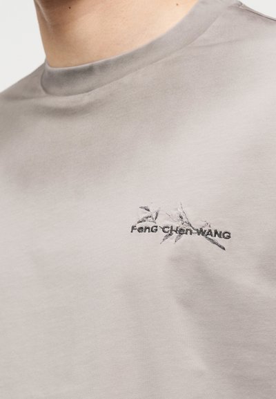 Μπεζ μπλούζα με στρογγυλή λαιμόκοψη, μικρό μαύρο κεντητό κλαδί και λογότυπο με κείμενο στην αριστερή πλευρά του στήθους.