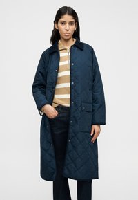 Μακρύ καπιτονέ παλτό σε ναυτικό μπλε χρώμα με γιακά, πλαϊνές τσέπες και κουμπιά με κλιπ, συνδυασμένο με ριγέ πουλόβερ και σκούρο τζιν. Λείο υλικό.