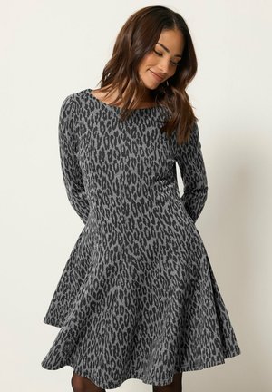 Mujer con cabello largo y castaño que lleva un vestido de manga larga de estampado de leopardo en gris y negro con una falda acampanada, de pie frente a un fondo claro y liso.
