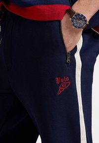 Tmavě modré tepláky s červeným logem "Polo", bílými bočními pruhy a kapsou na zip. Materiál působí texturovaně a odolně.