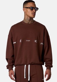 Brown sweatshirt with white logos, relaxed fit, crew neck, ribbed cuffs, and hem. Paired with matching pants featuring a white drawstring.