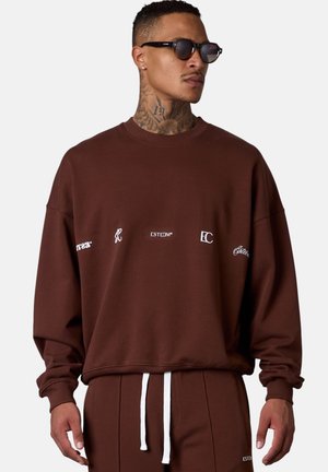 Brown sweatshirt with white logos, relaxed fit, crew neck, ribbed cuffs, and hem. Paired with matching pants featuring a white drawstring.